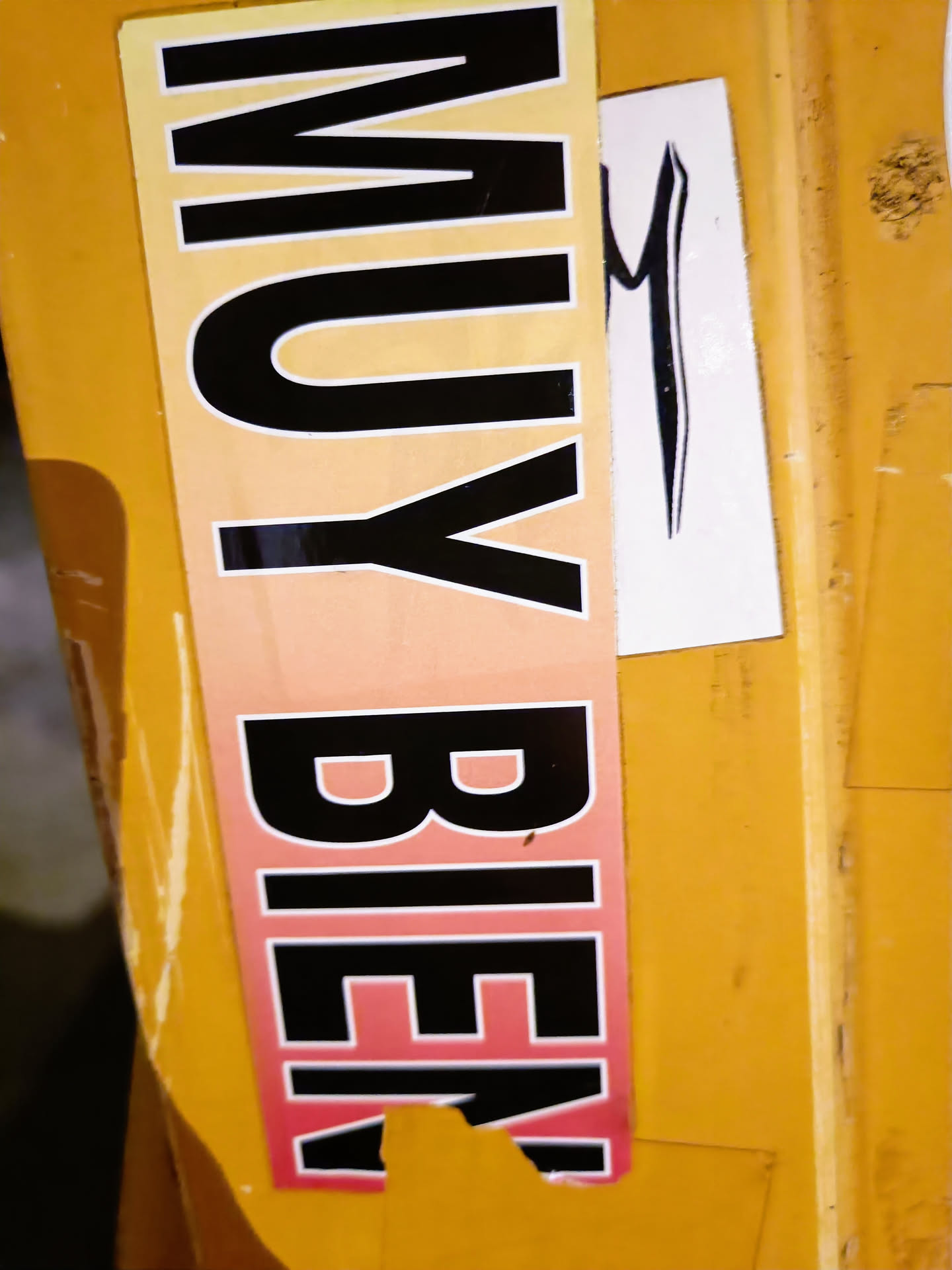 A sticker with the words "MUY BIEN" in black letters on a gradient orange-pink background.  The sticker is slightly damaged at the bottom. There is a smaller, separate sticker with a stylized black 'M' next to it.