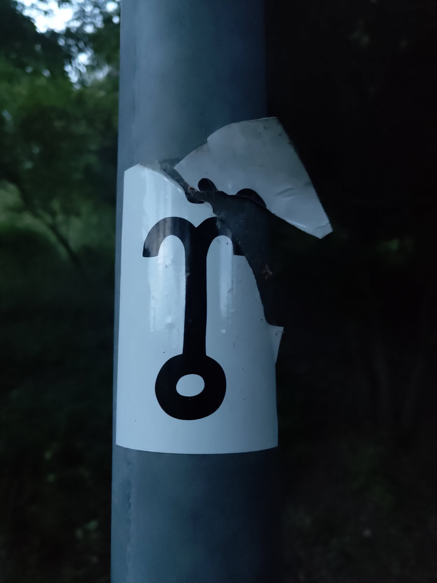 A white sticker affixed to a grey pole shows a black symbol that looks like a stylized pendulum or a simple abstract design.  Part of the sticker is torn away.
