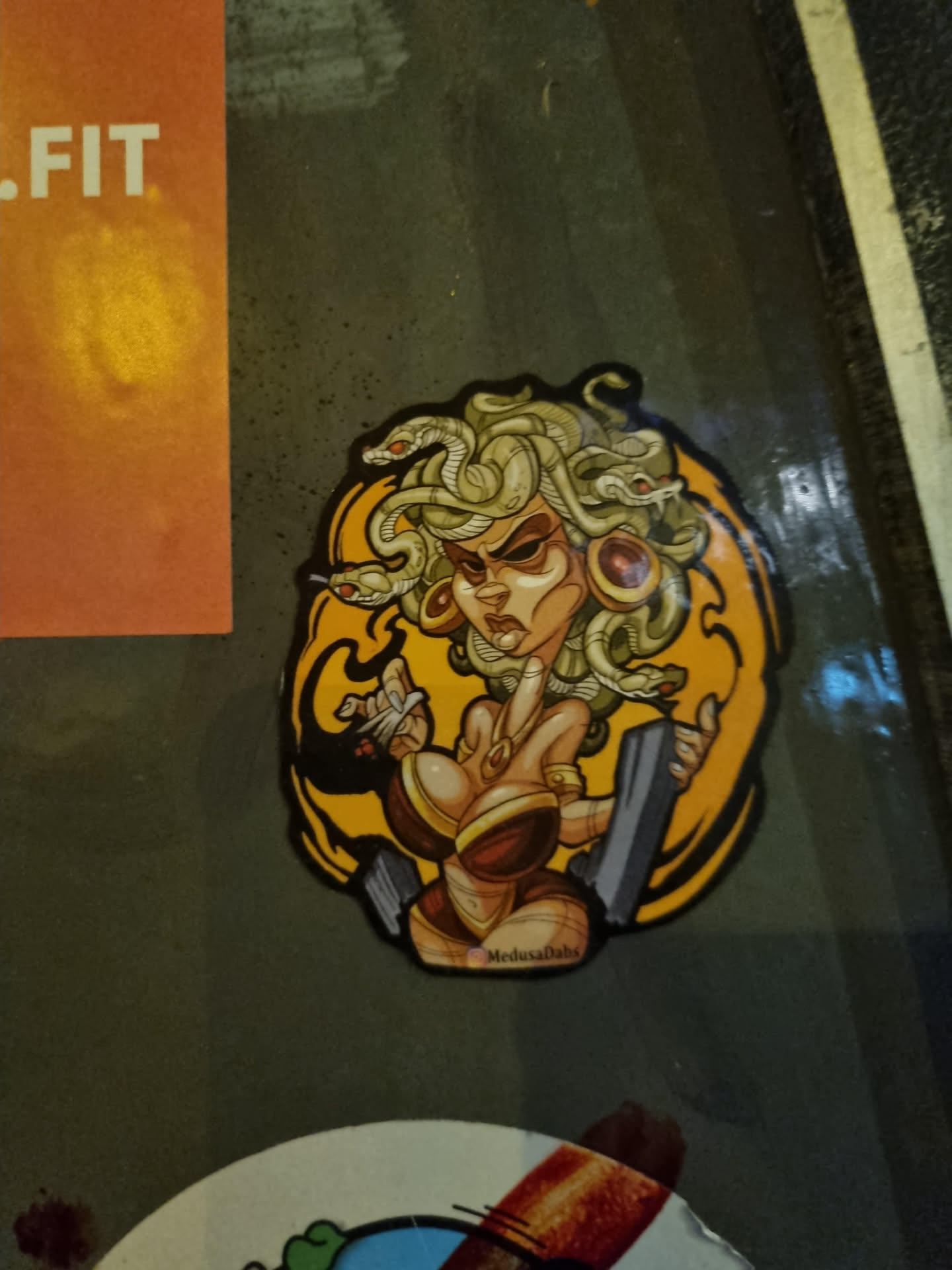 A sticker featuring a stylized illustration of Medusa, the Gorgon from Greek mythology. She is depicted as a voluptuous woman with snakes for hair, wearing a warrior-like outfit. The art style is cartoonish and vibrant, with a bold color palette. The sticker is round with a yellow background. It has the artist's signature at the bottom.