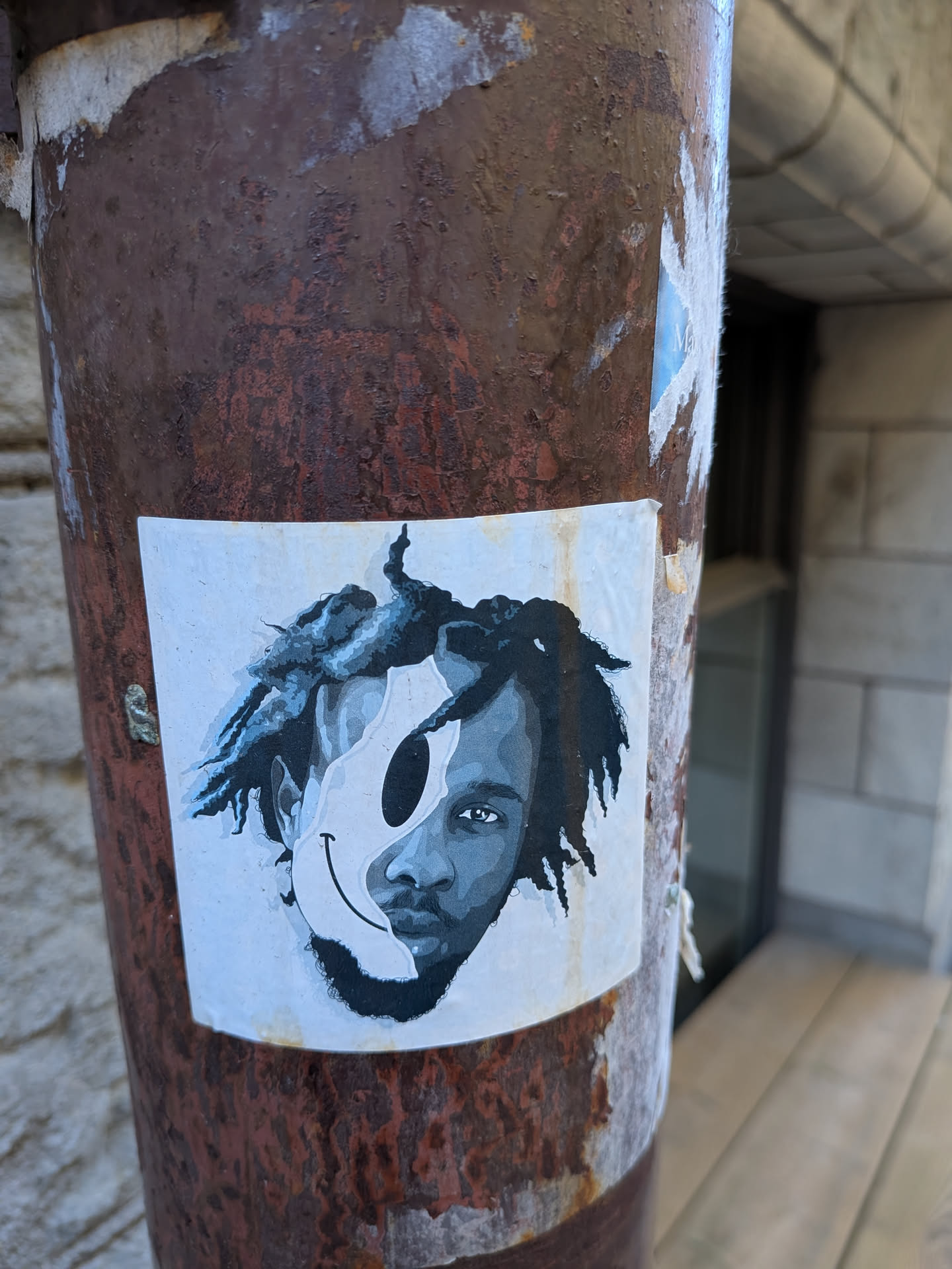 The sticker is a rectangular piece of paper featuring a monochromatic graphic. The left half of the sticker depicts the left side of a person's face, including an eye, nose, and mouth, rendered in shades of gray and black. This realistic portrait style contrasts with the right half, which is dominated by a simplified, somewhat distorted smiley face. The smiley face is black with white highlights and its mouth curves upwards. The overall effect is a fusion of a human portrait and an abstract symbol, creating a sense of duality. The sticker appears to be affixed to a rusty metal pole and shows some signs of wear, with slightly curled edges and a faint yellowing of the paper in some areas.