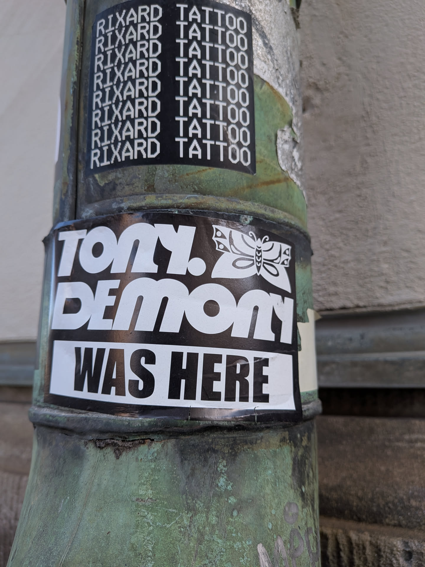 A rectangular vinyl sticker adhered horizontally around a weathered green metal pipe. The design is split into two sections: the top has a glossy black background featuring bold, stylized white text reading 'TONY. DEMON' accompanied by an ornate, tattoo-style butterfly graphic to the right of 'TONY.'. Below this is a white rectangular banner with bold black lettering stating 'WAS HERE'.