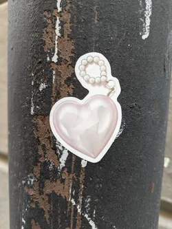 A cute sticker of a pink heart with a pearl charm attached. The sticker is on a dark gray textured surface.
