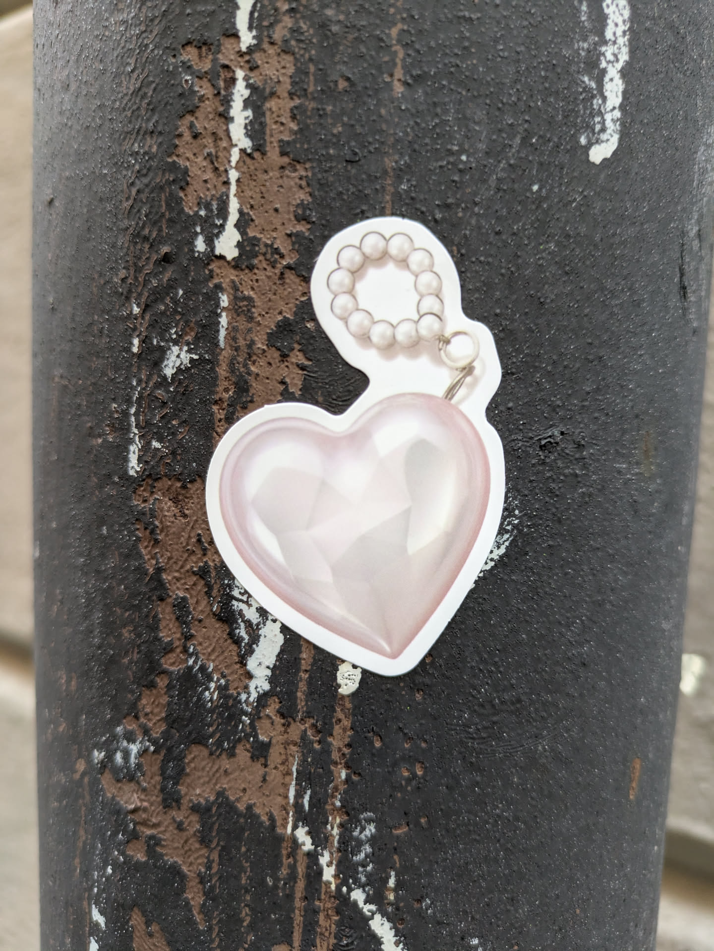 A cute sticker of a pink heart with a pearl charm attached. The sticker is on a dark gray textured surface.