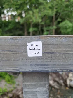 A white square sticker with the text "MIA MAGIA .COM" on a gray wooden surface. The background is blurred, showing green trees.
