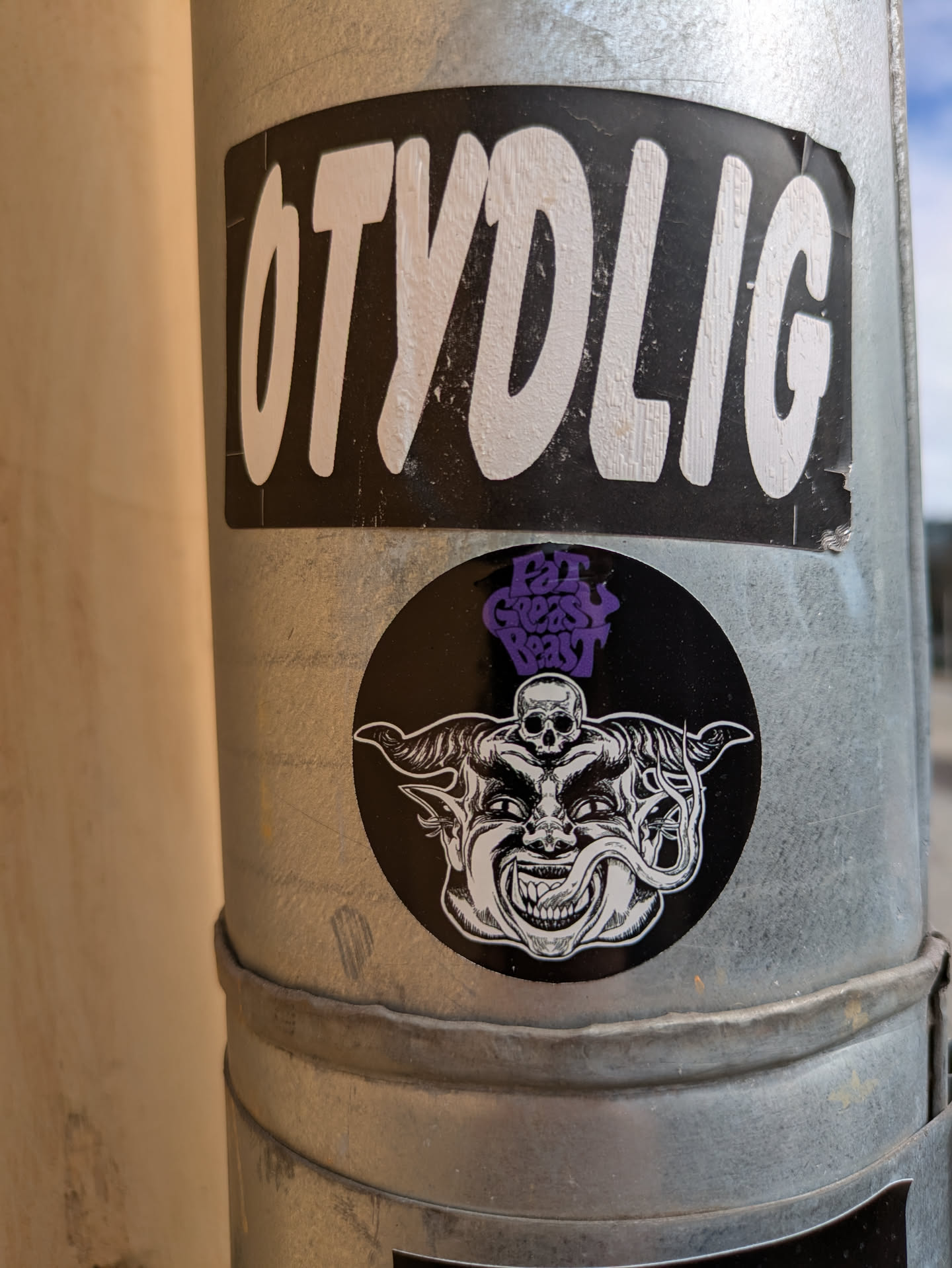 A round sticker showing a black and white drawing of a demon face with horns and a skull on its head, stuck to a light pole. The sticker at the top reads "Eat Grease Reat".
