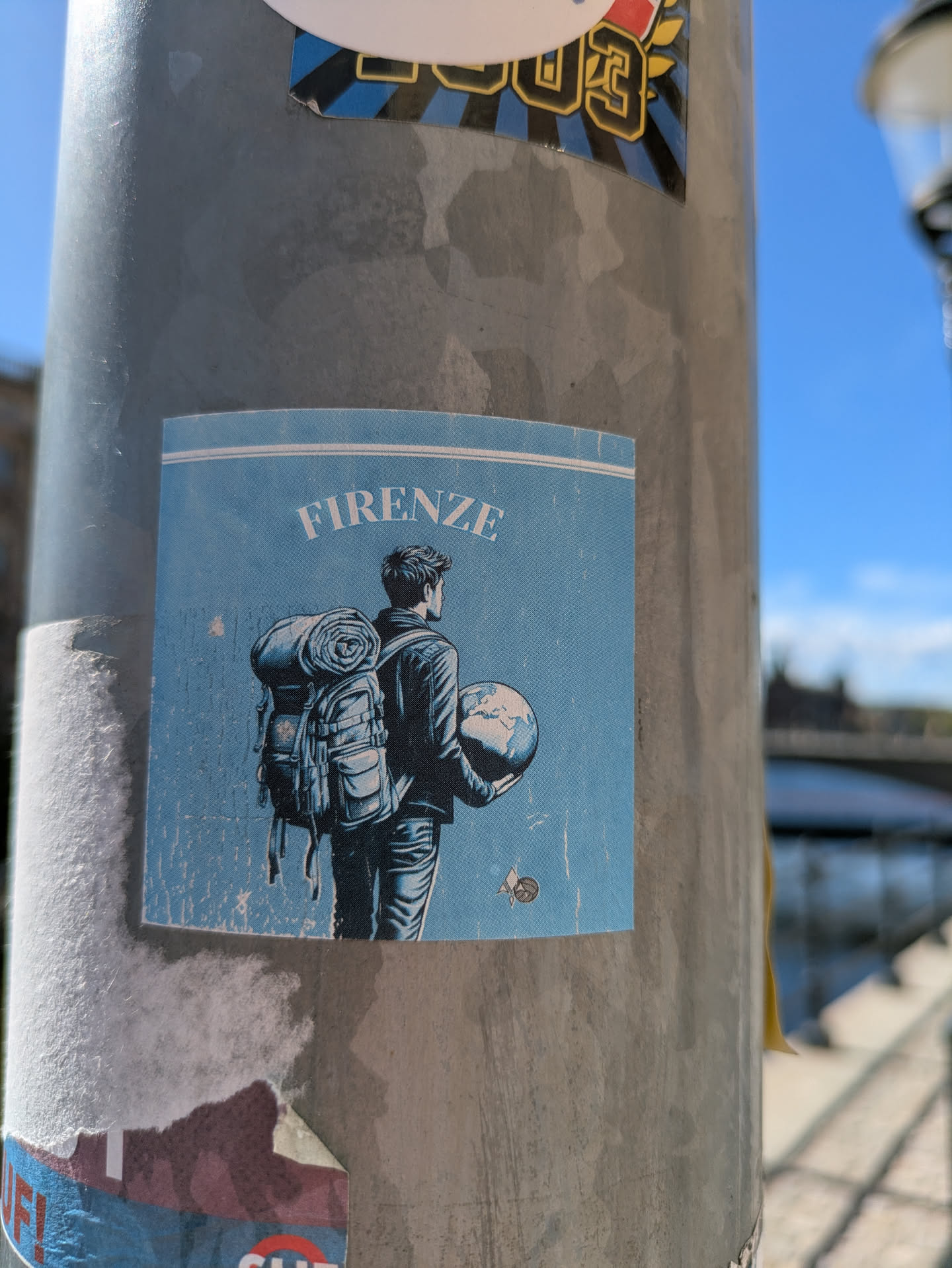 A square sticker is adhered to a gray pole, featuring a traveler with a backpack and holding a globe, with the word "FIRENZE" printed at the top. The sticker has a blue-ish color palette.