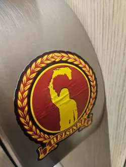 A circular sticker featuring a bold design in dark red, yellow, and black. The central motif is a dark red circle bordered by a vibrant yellow laurel wreath. Inside the red circle, a yellow silhouette of a person is depicted from the chest up, holding an irregularly shaped yellow object above their head, resembling a stylized map of Finland or a flame. Below the central circle, a yellow banner with a black outline unfurls, bearing the word 'HELSINKI' in black capital letters. The sticker shows visible creases and wrinkles, indicating it is a physical sticker applied to a brushed metal surface.