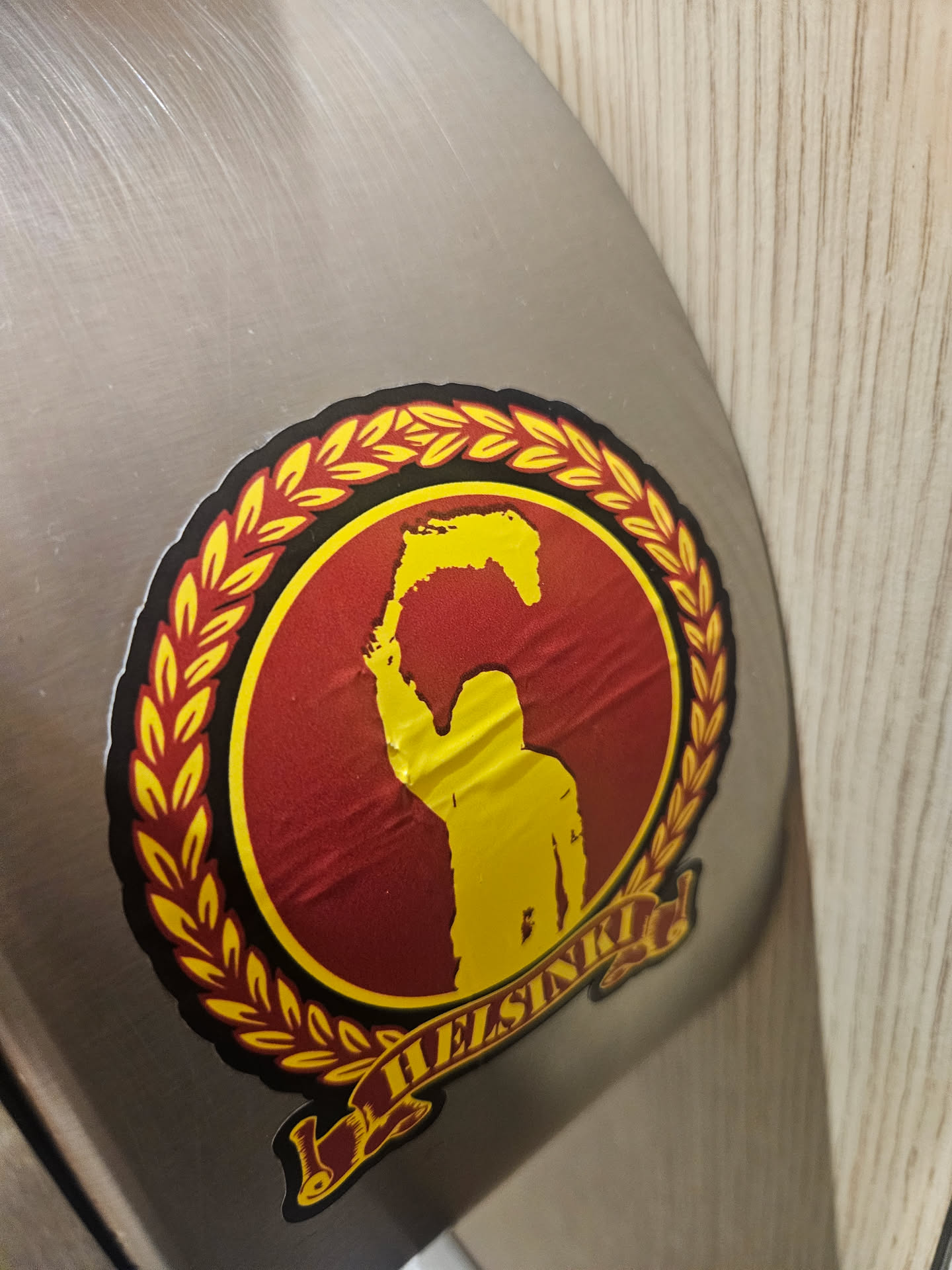 A circular sticker featuring a bold design in dark red, yellow, and black. The central motif is a dark red circle bordered by a vibrant yellow laurel wreath. Inside the red circle, a yellow silhouette of a person is depicted from the chest up, holding an irregularly shaped yellow object above their head, resembling a stylized map of Finland or a flame. Below the central circle, a yellow banner with a black outline unfurls, bearing the word 'HELSINKI' in black capital letters. The sticker shows visible creases and wrinkles, indicating it is a physical sticker applied to a brushed metal surface.