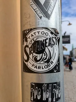 A black and white sticker on a pole advertises "Speakeasy Tattoo Parlour" in a stylized design. The sticker is torn at the top and is placed above another sticker that reads "The Hell Tee"