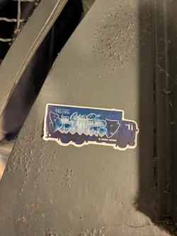 Street sticker A die-cut sticker shaped like a delivery box truck facing right. The truck is colored dark blue and overlaid with stylized blue-and-white graffiti lettering across the cargo container. Above the large lettering is a cursive logo reminiscent of the Coca-Cola script, and small tag signatures appear in the corners, including an Instagram handle near the bottom right rear wheel.