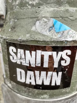 A black sticker with white text that says "SANITYS DAWN" on a weathered surface. The sticker shows some signs of wear and tear.