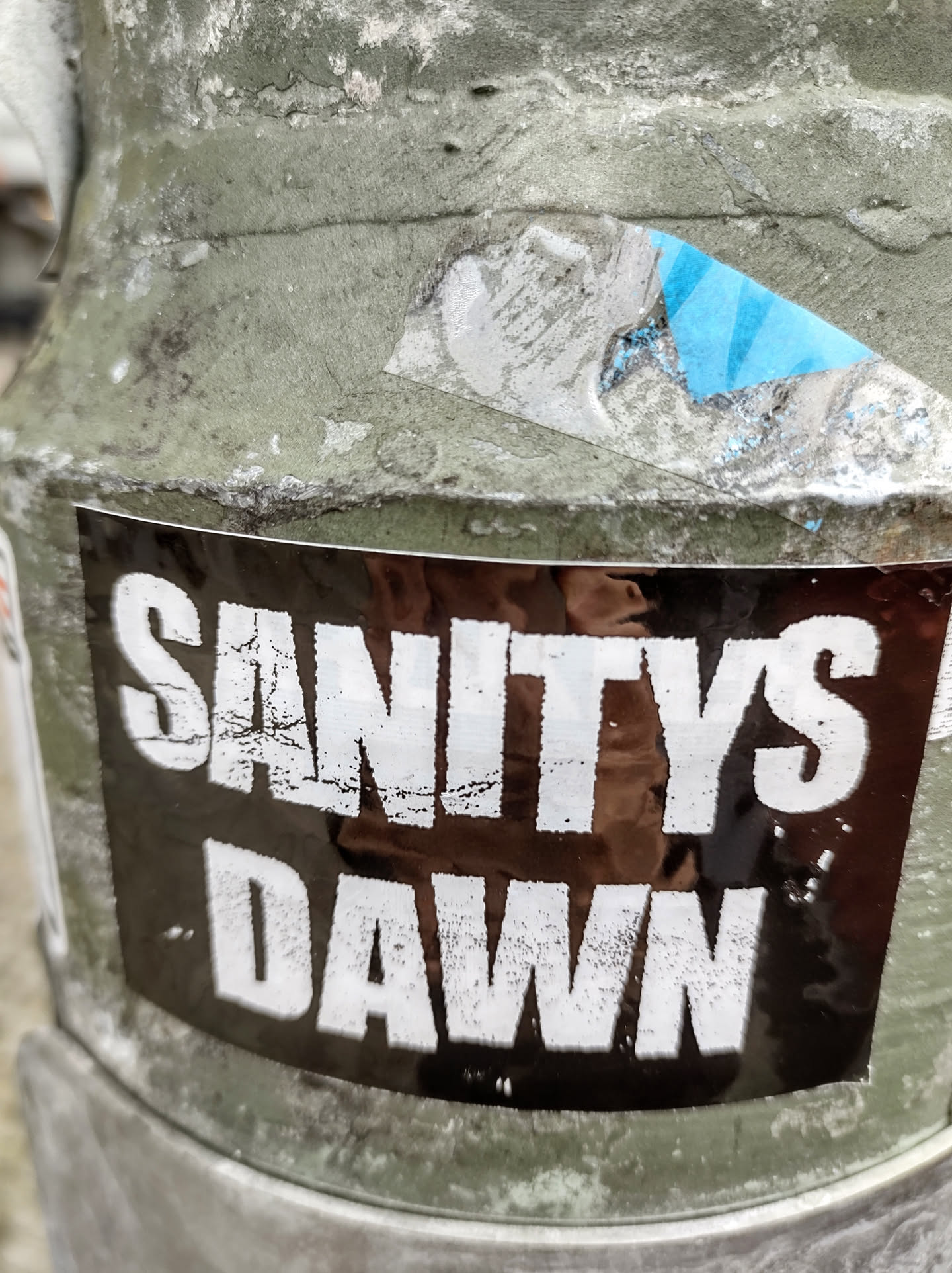 A black sticker with white text that says "SANITYS DAWN" on a weathered surface. The sticker shows some signs of wear and tear.