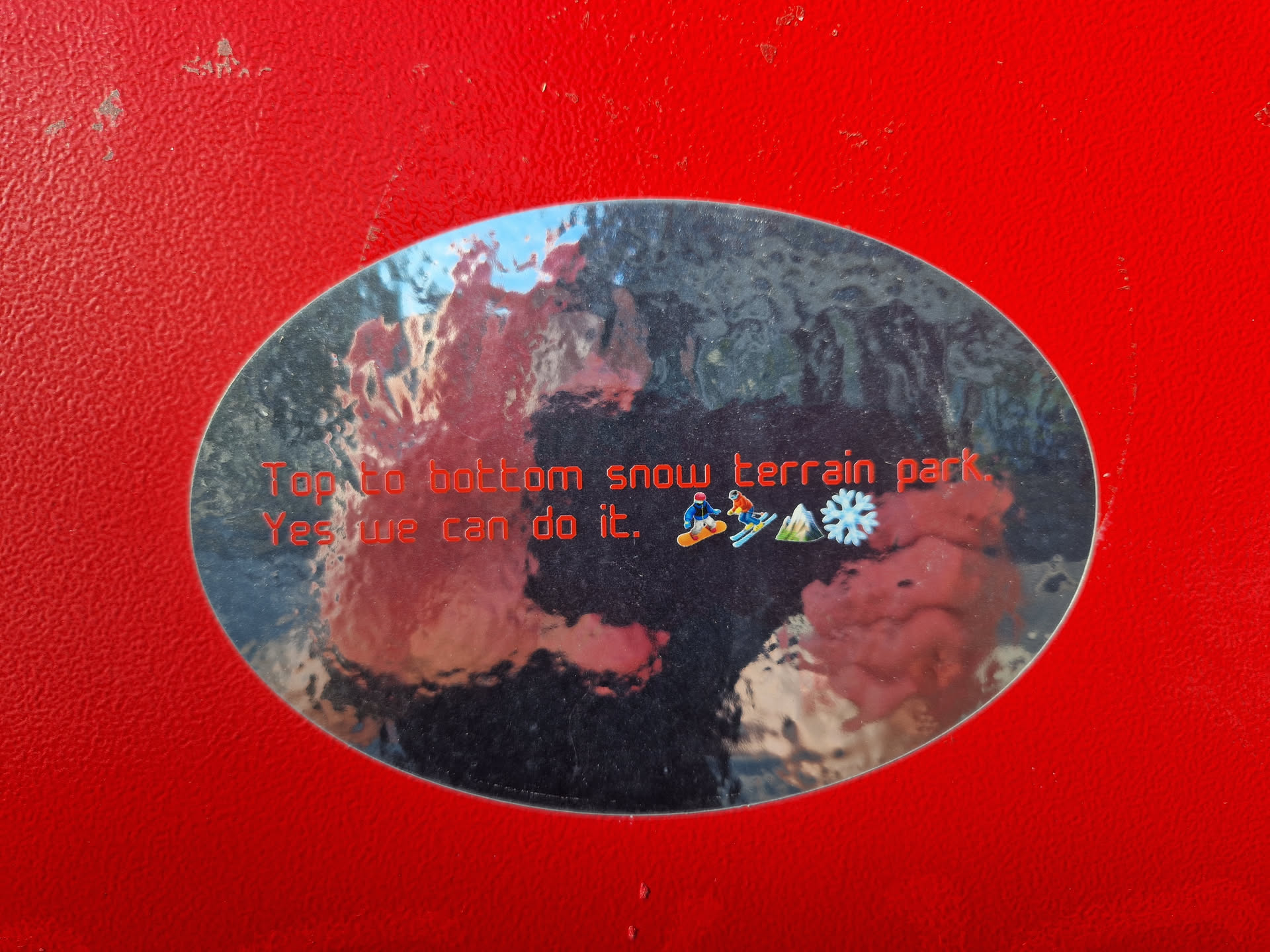 Oval-shaped sticker with a blurry background image and the text 'Top to bottom snow terrain park. Yes we can do it.' in red.  Small icons of skiers and snowboarders are present as well.