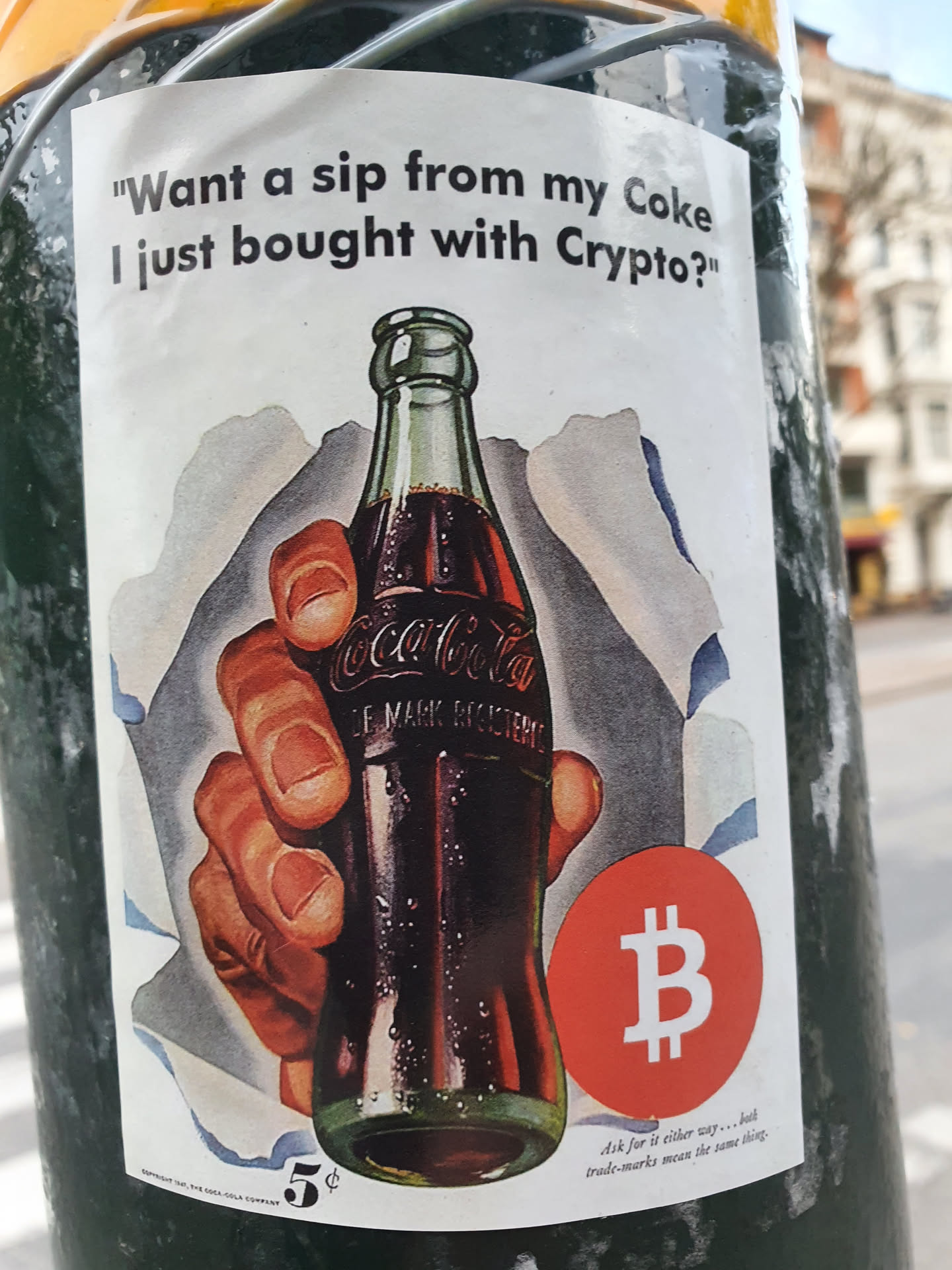 A sticker featuring a vintage Coca-Cola advertisement with a Bitcoin symbol overlaid. The text on the sticker asks if you want a sip of Coke bought with crypto.
