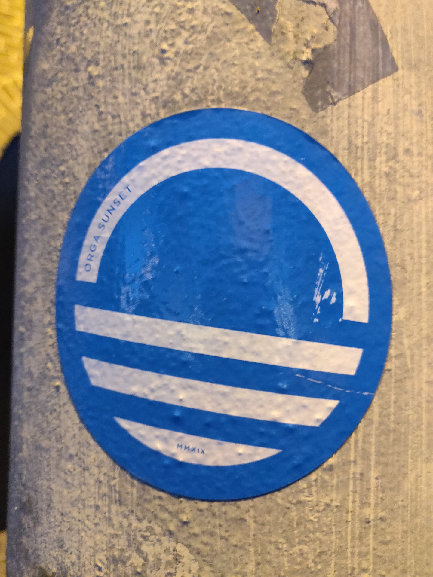 Round blue sticker with a white design. The text 'ORGASUNSET' is visible on the left side and 'HMAXII' at the bottom. The design is simple and modern, featuring white concentric circles and lines on a blue background.