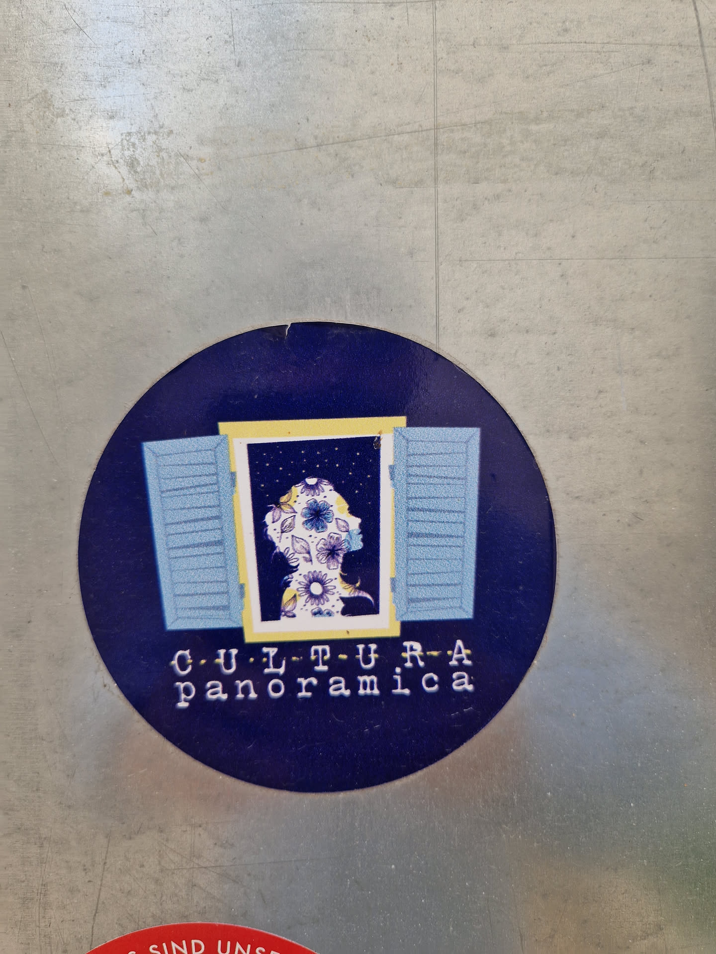 Round sticker featuring a silhouette of a person's head and shoulders adorned with flowers, seen through a light blue window. The background is dark blue, and the text 'CULTURA Panoramica' is written in a typewriter font below the image.