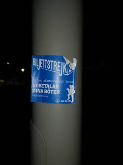 The sticker is a rectangular, blue-backed decal with white text and graphics, affixed to a grey pole. The top right corner is slightly peeled and torn, showing some wear. The prominent text at the top reads "BILJETTSTREJK" (Ticket Strike). Below this, partially obscured, is "Ak med rabatterad biljett i januari" (Ride with discounted ticket in January). The central message states "VI BETALAR DINA BÖTER" (We pay your fines). To the right, a white silhouette depicts two stylized figures, one larger and one smaller, possibly representing solidarity or a family. At the bottom left, the website "biljettstrejk.se" is visible, and below the figures, a small circular logo contains the text "Allt åt alla" (Everything for everyone). The overall aesthetic is simple, direct, and activist-oriented.