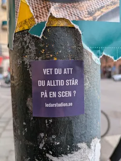 A blue sticker on a weathered pole with text in Swedish, that says, "Do you know that you are always on a stage?". The sticker also has a website address.