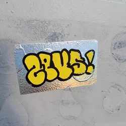 A silver holographic sticker with yellow graffiti-style lettering that says "GLUE!" in bubble letters. The sticker is attached to a light gray wall with some texture.