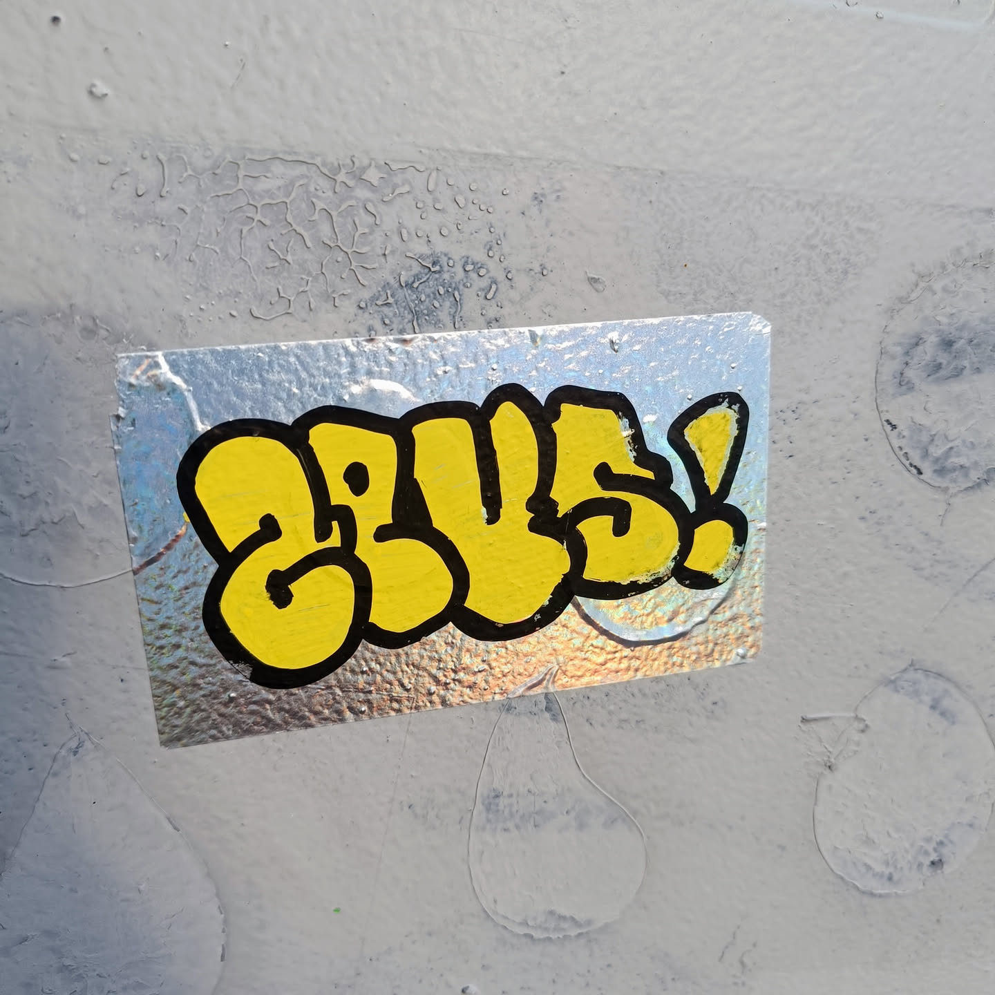 A silver holographic sticker with yellow graffiti-style lettering that says "GLUE!" in bubble letters. The sticker is attached to a light gray wall with some texture.