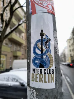 A sticker featuring a stylized blue snake wrapped around the Berlin Television Tower.  The text 'INTER CLUB BERLIN' is prominently displayed below the image. The sticker appears to be affixed to a street pole.