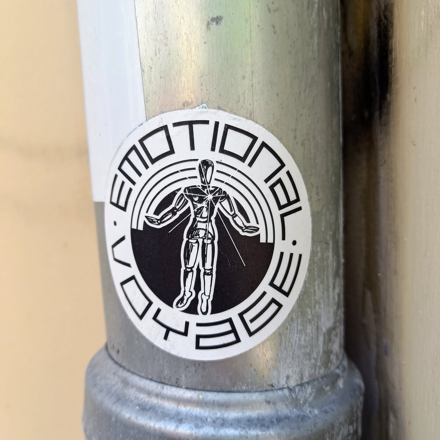 Black and white circular sticker featuring a robotic figure at the center with the words "Emotional Voyage" written around it. The figure has its arms outstretched, and appears to be radiating light. The text is stylized in a geometric font.