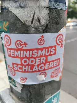 A sticker expressing a choice between feminism and violence. It features the text "FEMINISMUS ODER SCHLÄGEREI" (Feminism or Violence) in red on a white background, along with several feminist symbols. The sticker is affixed to a pole, surrounded by other stickers and debris.