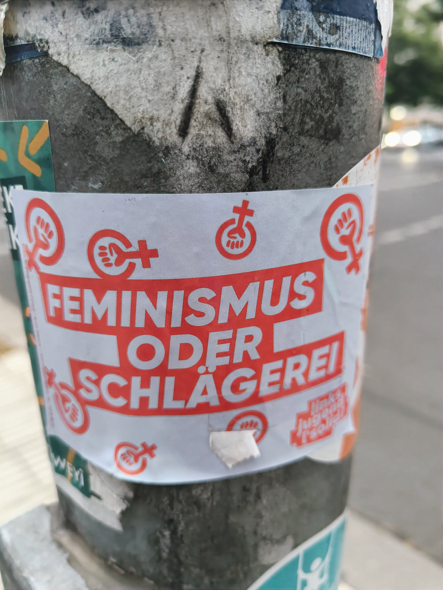 A sticker expressing a choice between feminism and violence. It features the text "FEMINISMUS ODER SCHLÄGEREI" (Feminism or Violence) in red on a white background, along with several feminist symbols. The sticker is affixed to a pole, surrounded by other stickers and debris.