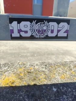 A rectangular sticker with a black border, featuring a design of bold white numbers '19' on the left and '02' on the right. In the center, between the numbers, is a white emblem consisting of a laurel wreath surrounding intertwined letters, which appear to be 'C' and 'A' or 'A' and 'C'. The background of the sticker, including the fill of the central emblem's letters, is horizontally striped in alternating purple and white bands. The sticker is adhered to a grey metal beam.