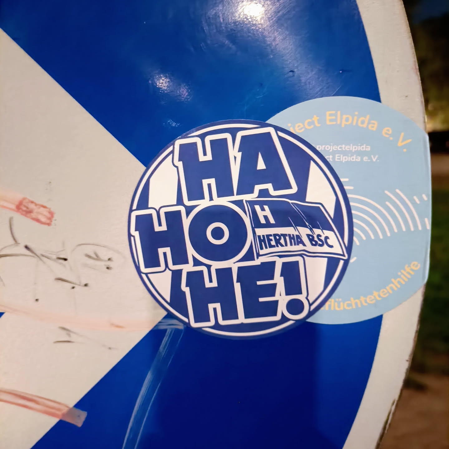 A round sticker with the text "Ha Ho He!" in blue on a white background.  There is a small Hertha BSC logo included in the design. The sticker is affixed to a blue surface.