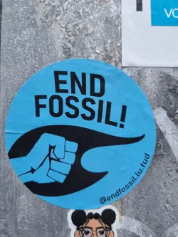 A round blue sticker with a black fist and the text END FOSSIL!. The sticker also contains a social media handle and a website address.