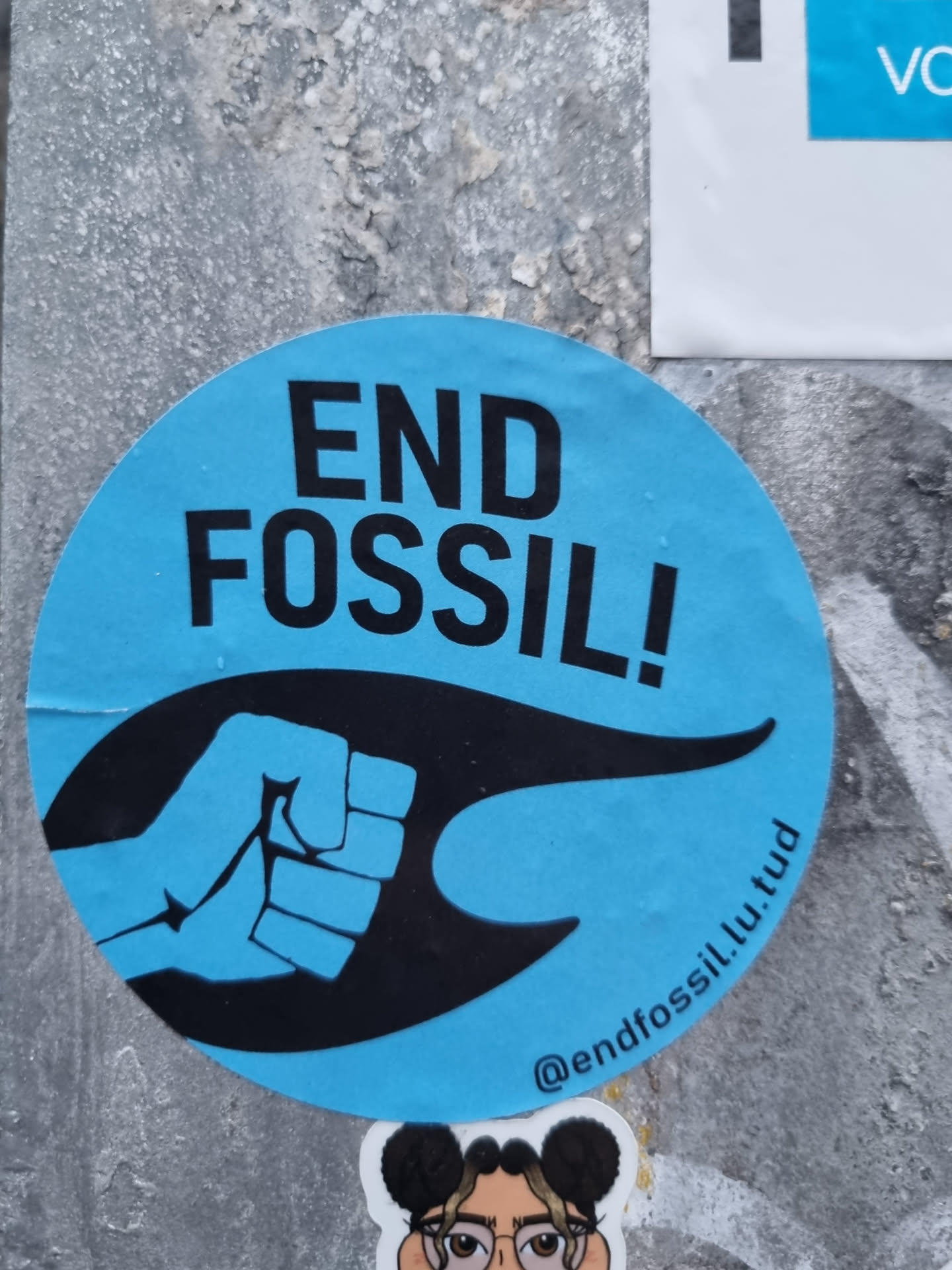 A round blue sticker with a black fist and the text END FOSSIL!. The sticker also contains a social media handle and a website address.