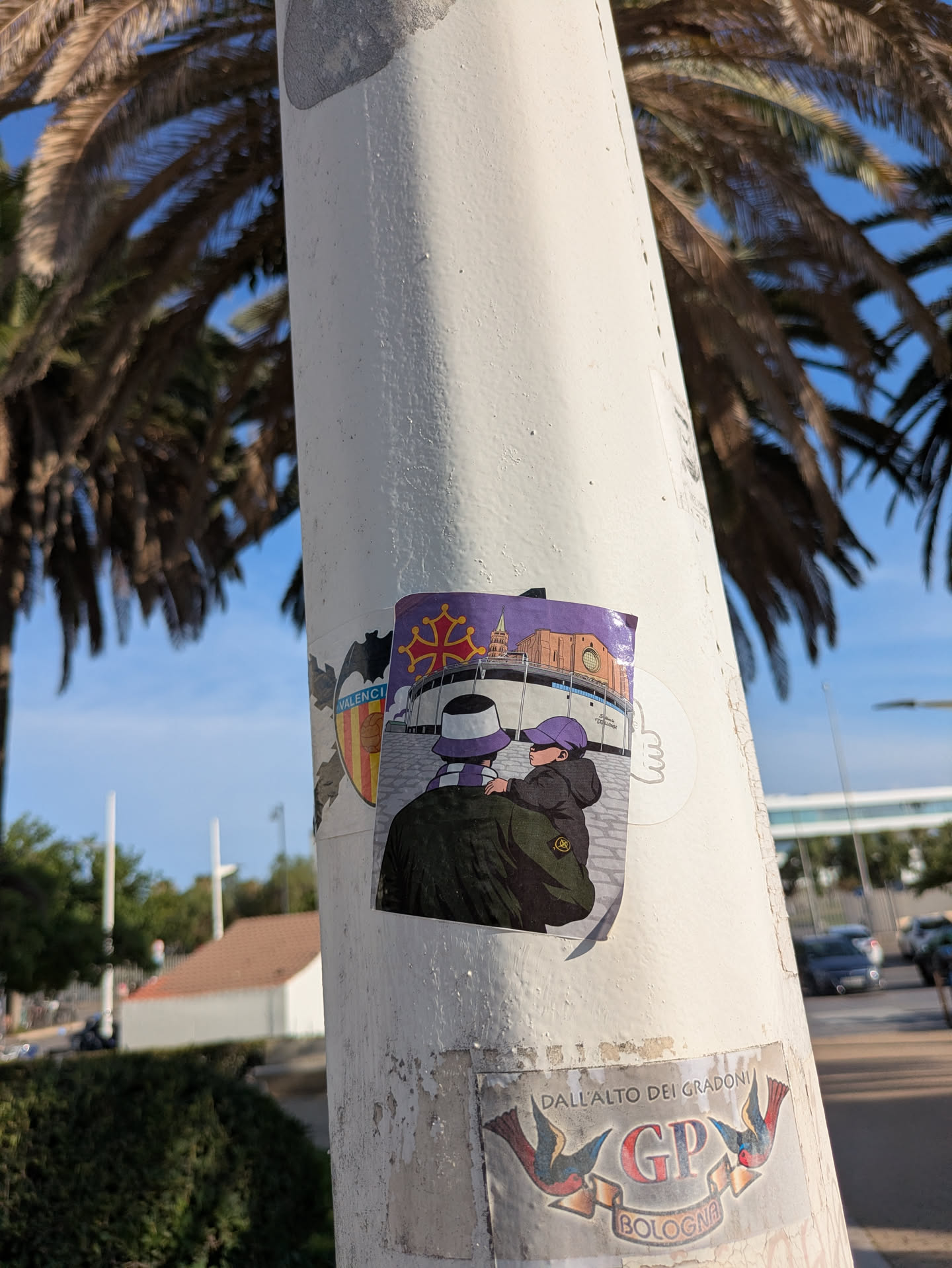 This sticker features two individuals, one in a black jacket with a Stone Island patch and a purple cap, and another in a black jacket, a purple and white scarf, and a purple hat. They appear to be standing in front of a stadium, with a purple sky overhead. The stadium has a symbol in the sky with a tower on top, suggesting it might be related to a specific location or team.
