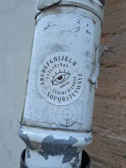 Street sticker A circular sticker affixed to a weathered white pipe, showcasing a design inspired by a spirit board. At the center is a stylized open human eye with extended eyelashes. Above the eye arcs the sequence of numbers '1234567890', enveloped along the outer edge by the letters 'A B C D E F G H I J K L M'. Directly beneath the eye reads 'GOOD BYE' and 'VASUMA EYEWEAR', bordered on the lower circumference by the remaining letters 'N O P Q R S T U V W X Y Z'. The print is rendered in a dark sepia or black ink on an off-white circular background.