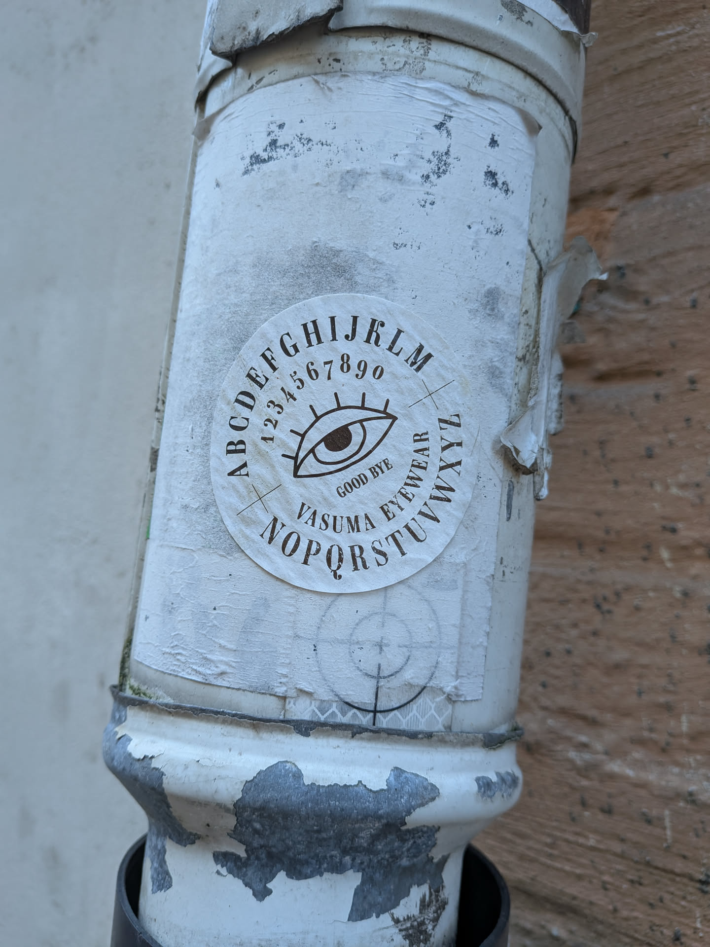 A circular sticker affixed to a weathered white pipe, showcasing a design inspired by a spirit board. At the center is a stylized open human eye with extended eyelashes. Above the eye arcs the sequence of numbers '1234567890', enveloped along the outer edge by the letters 'A B C D E F G H I J K L M'. Directly beneath the eye reads 'GOOD BYE' and 'VASUMA EYEWEAR', bordered on the lower circumference by the remaining letters 'N O P Q R S T U V W X Y Z'. The print is rendered in a dark sepia or black ink on an off-white circular background.