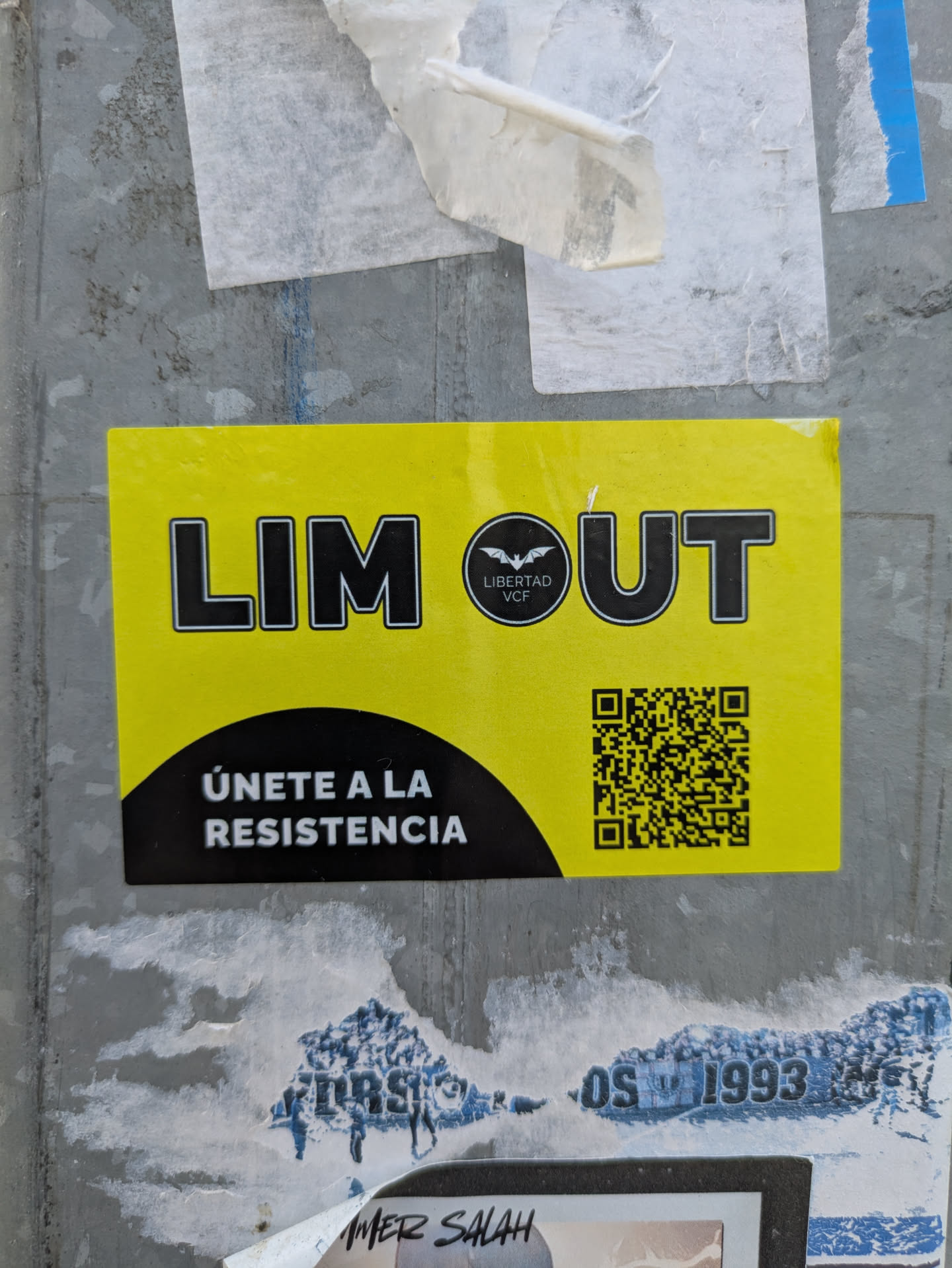 A bright yellow sticker affixed to a weathered gray surface, partially covered by remnants of other stickers. The sticker prominently displays the text "LIM OUT" in bold, black lettering. A circular emblem featuring a bat and the words "LIBERTAD VCF" is positioned between the two words. Below this, the phrase "ÚNETE A LA RESISTENCIA" (Join the Resistance) is written in a similar black font, followed by a QR code.