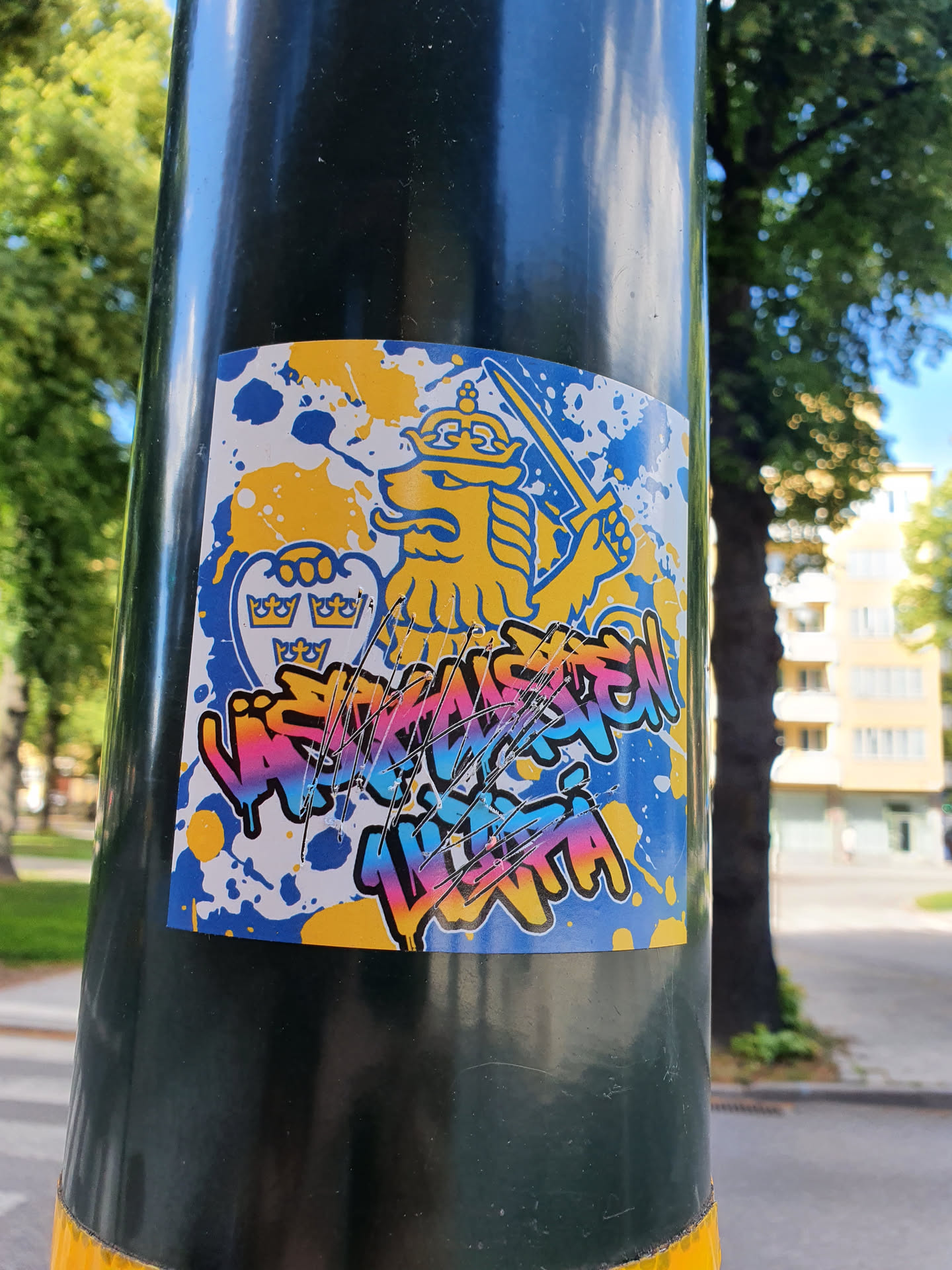 A colorful sticker featuring the Swedish coat of arms lion in a graffiti style. The background is a mix of blue and yellow splotches. The text below the lion appears to say "VÄSTERMÄSTEN LILLA VITA", though it is somewhat damaged.