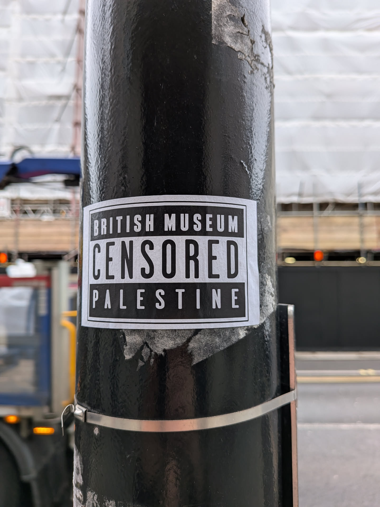 A rectangular, black and white sticker affixed to a textured black pole. The sticker features a black background with a thin white border. It contains three lines of white, sans-serif, uppercase text. The top line reads 'BRITISH MUSEUM', the middle line, which is significantly larger and bolder, reads 'CENSORED', and the bottom line reads 'PALESTINE'. The sticker shows minor signs of wear, with slight peeling visible at the edges.