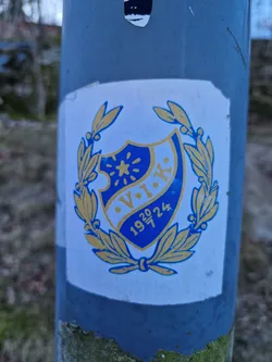 A sticker featuring a blue and gold crest with the letters V.I.K. and the numbers 1920-24.  It's encased within a laurel wreath. The style is reminiscent of vintage club or society emblems.