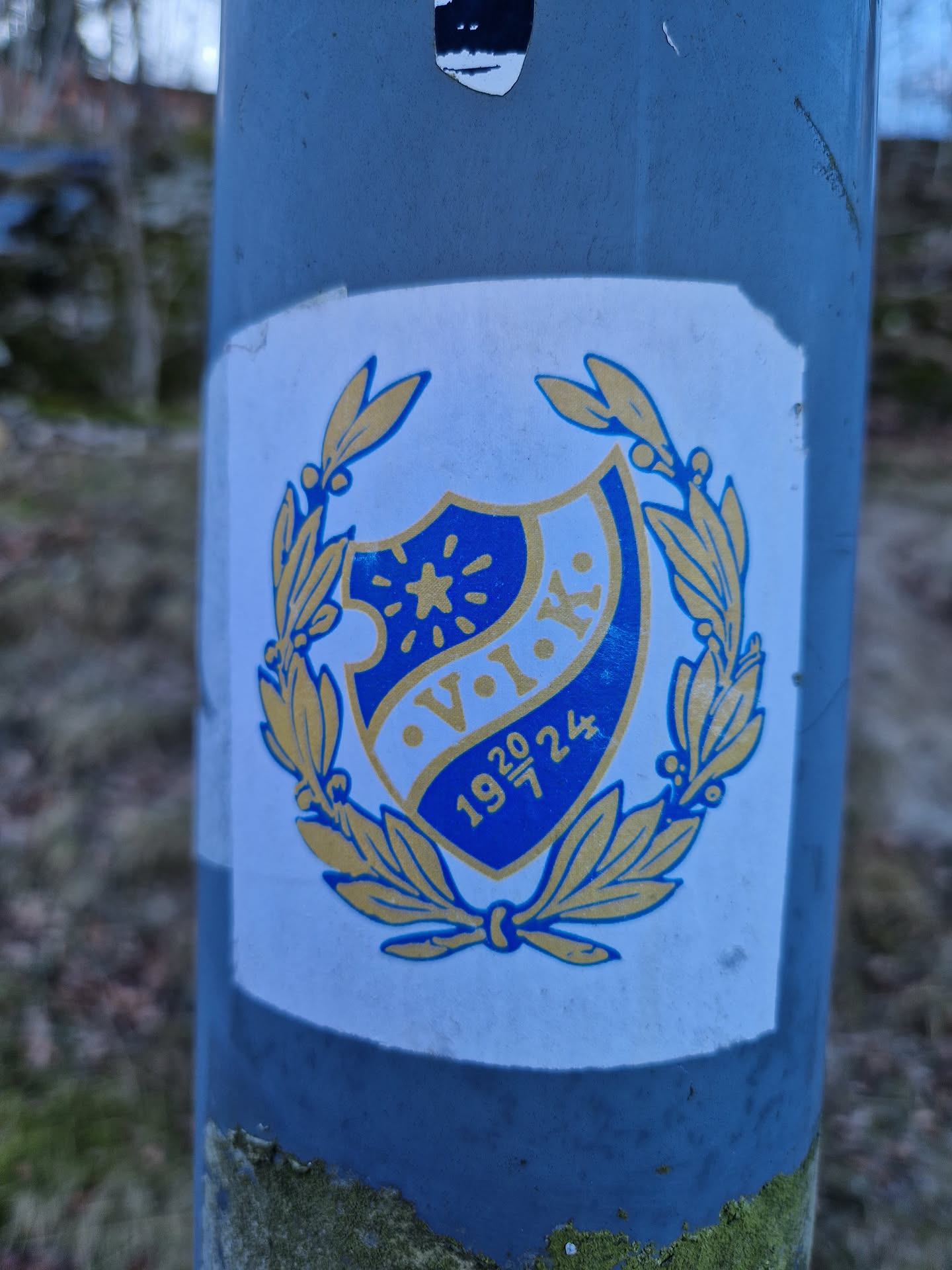 A sticker featuring a blue and gold crest with the letters V.I.K. and the numbers 1920-24.  It's encased within a laurel wreath. The style is reminiscent of vintage club or society emblems.