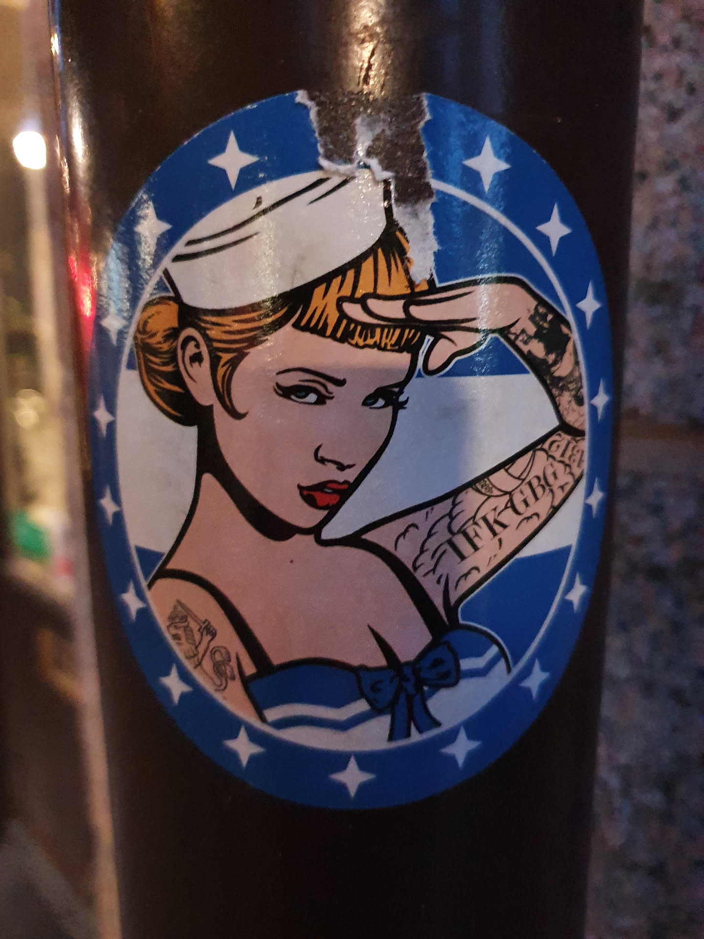 A round sticker featuring a pin-up illustration of a sailor girl with tattoos saluting. The design is in a vintage style with a blue and white color scheme and stars around the border.  The sticker shows some signs of wear and tear.