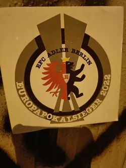 A sticker commemorating EFC Adler Berlin's victory in the Europapokal in 2022. The sticker features a circular design with the team's logo, which includes a red eagle and a black bear, symbolizing the team's name (Adler means eagle in German). The text around the logo indicates the team's name and the year of their victory. The overall design is in shades of gray and beige, giving it a vintage or classic feel.
