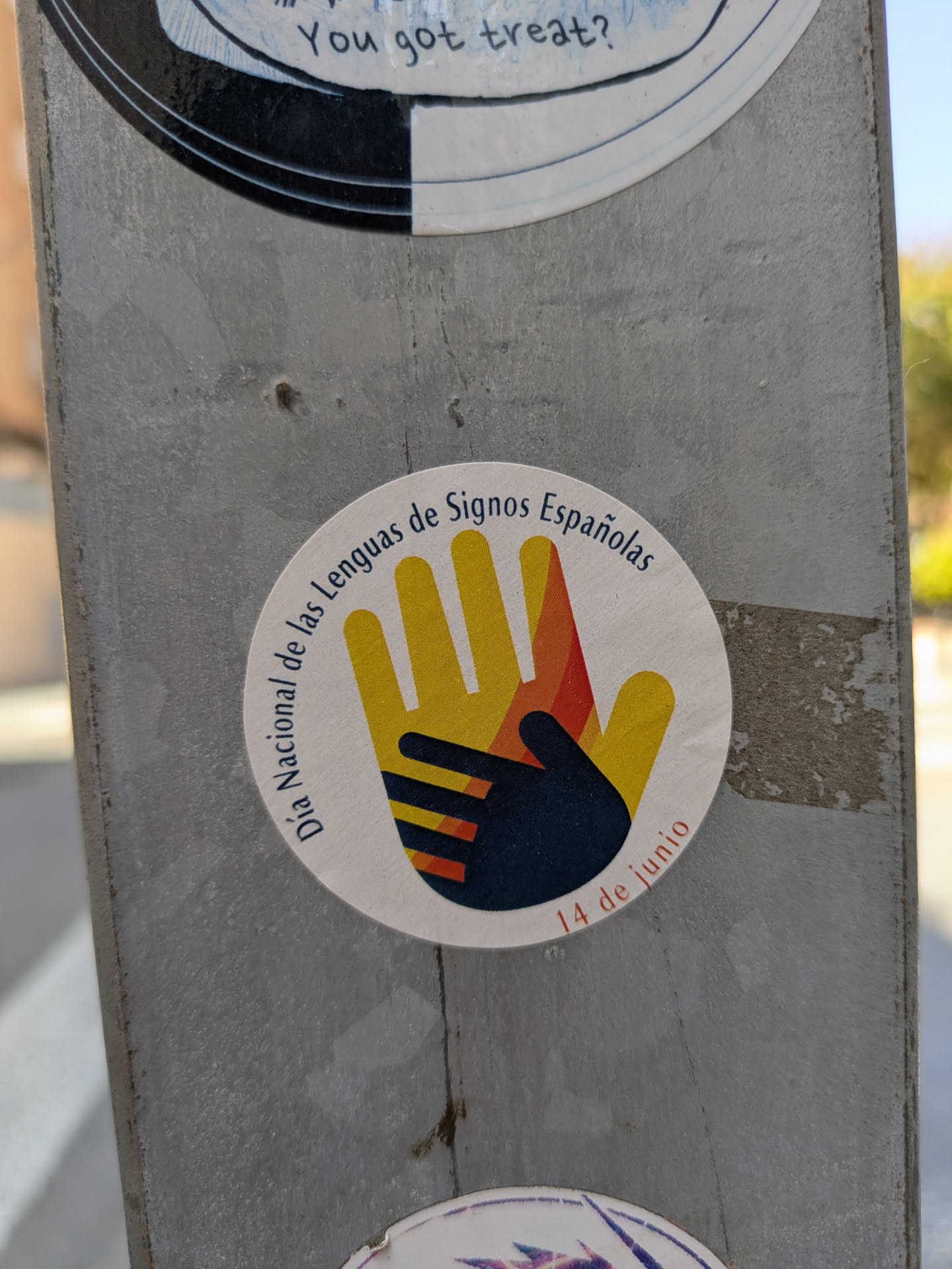 The sticker is round and placed on a metallic surface. It shows a large yellow and orange hand with a smaller dark hand inside. The text 'Día Nacional de las Lenguas de Signos Españolas' (National Day of Spanish Sign Languages) circles the top of the design, and '14 de junio' (June 14th) is written at the bottom.