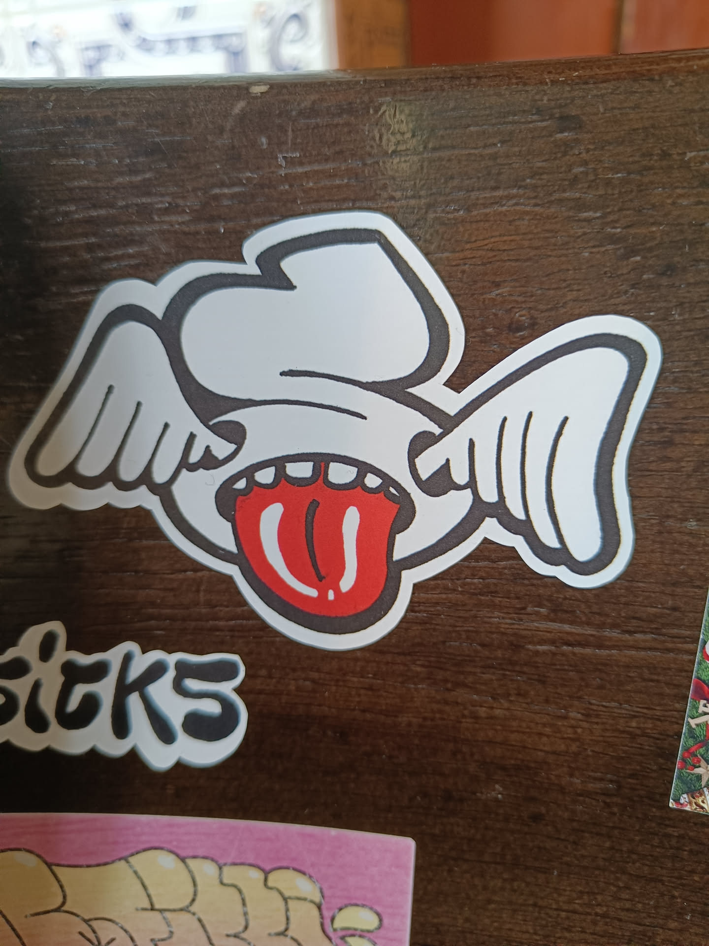 A sticker depicting a cartoonish face with wings and a long tongue sticking out. The style is playful and slightly edgy.