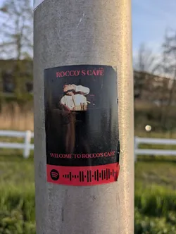 This is a sticker on a pole with a black background and an image of a person at a table, with the text "Rocco's Cafe" at the top, and "Welcome to Rocco's Cafe" at the bottom, along with a Spotify code.
