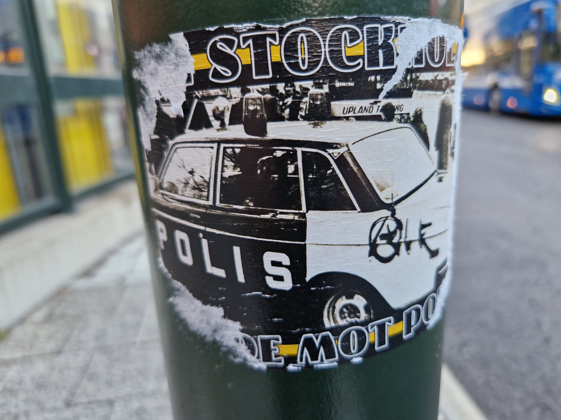 A sticker featuring a vintage police car, with text suggesting opposition to the police.  The sticker is affixed to a dark green pole, and appears worn and partially peeled.