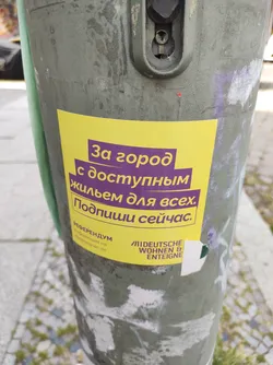 A yellow sticker affixed to a pole, promoting a referendum for affordable housing. The text is primarily in Russian, with additional text at the bottom in German, indicating a potential organization or initiative behind the campaign.