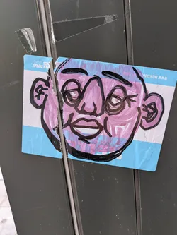 A sticker featuring a drawing of a face with a pink complexion, black outlines, and a light blue background. The sticker is slightly torn. 