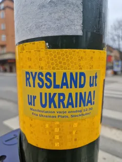 A yellow sticker on a pole with text in Swedish that translates to "Russia out of Ukraine!"  It also indicates a demonstration every Wednesday at 12:30 at Fria Ukrainas Plats in Stockholm.