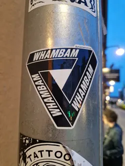 A black and white triangular sticker with the word WHAMBAM written in a repeating pattern. The sticker is affixed to a metal pole.  A small text is visible in the top right corner, presumably a social media handle.